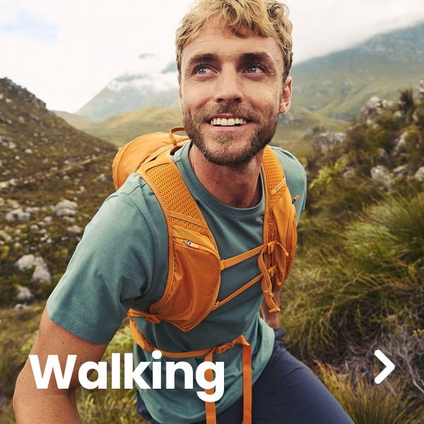 A person wearing a blue-green T-shirt and an orange hiking backpack walks along a rugged mountain trail surrounded by grassy hills and rocky terrain. Mountains rise in the background under a cloudy sky. Large white text reading “Walking” appears in the lower-left corner, and a right-arrow navigation icon is displayed on the right side of the image. The scene highlights outdoor walking and hiking in a scenic landscape.