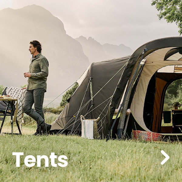 A large family-sized tent is pitched on a grassy campsite with mountains and misty skies in the background. A person stands beside outdoor camping furniture near the tent entrance, which is open to reveal part of the interior. Large white text reading “Tents” appears in the lower-left corner, and a right-arrow navigation icon is shown on the right side of the image. The scene is set outdoors in soft natural light.