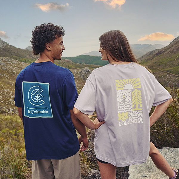 Two people stand on a rocky mountain hillside, viewed from behind, wearing oversized Columbia graphic T-shirts. One T-shirt is navy blue with a large blue-and-white logo graphic on the back, and the other is light grey with a yellow-and-white nature-inspired graphic. Rolling hills and mountains stretch into the background beneath a partly cloudy sky. The outdoor scene focuses on casual apparel in a scenic hiking landscape.