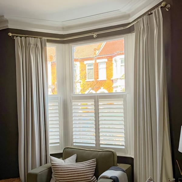 Bay window with white café style shutters, framed by tall cream curtains; soft daylight falls over an armchair in a cosy living room.