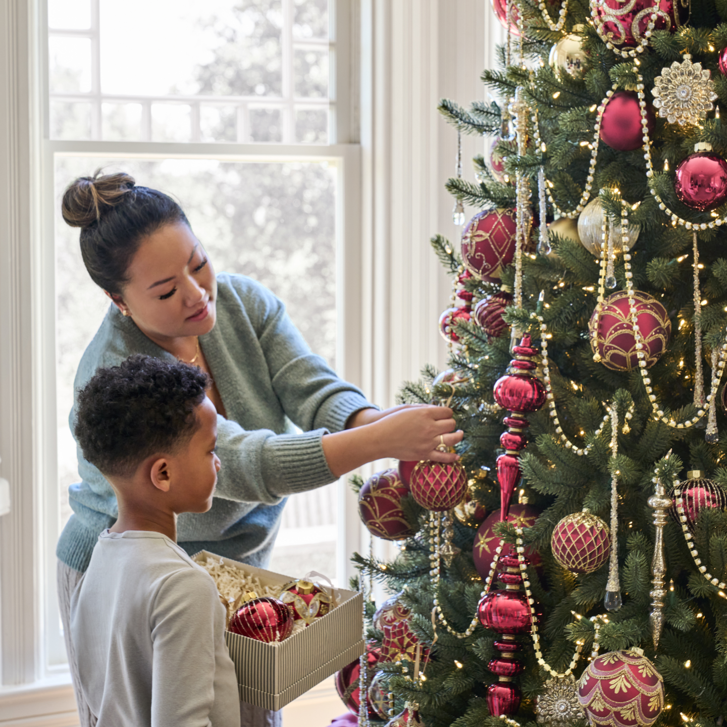 Woman and child decorating a Christmas tree with red and gold ornaments, beaded garlands, and warm lights.