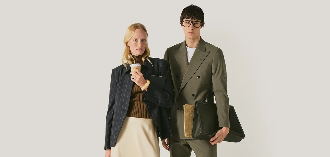 Uomo e donna in piedi, entrambi vestiti in abbigliamento smart casual; la donna tiene in mano una tazza di caffè e una borsa, l'uomo tiene una grande borsa di pelle.