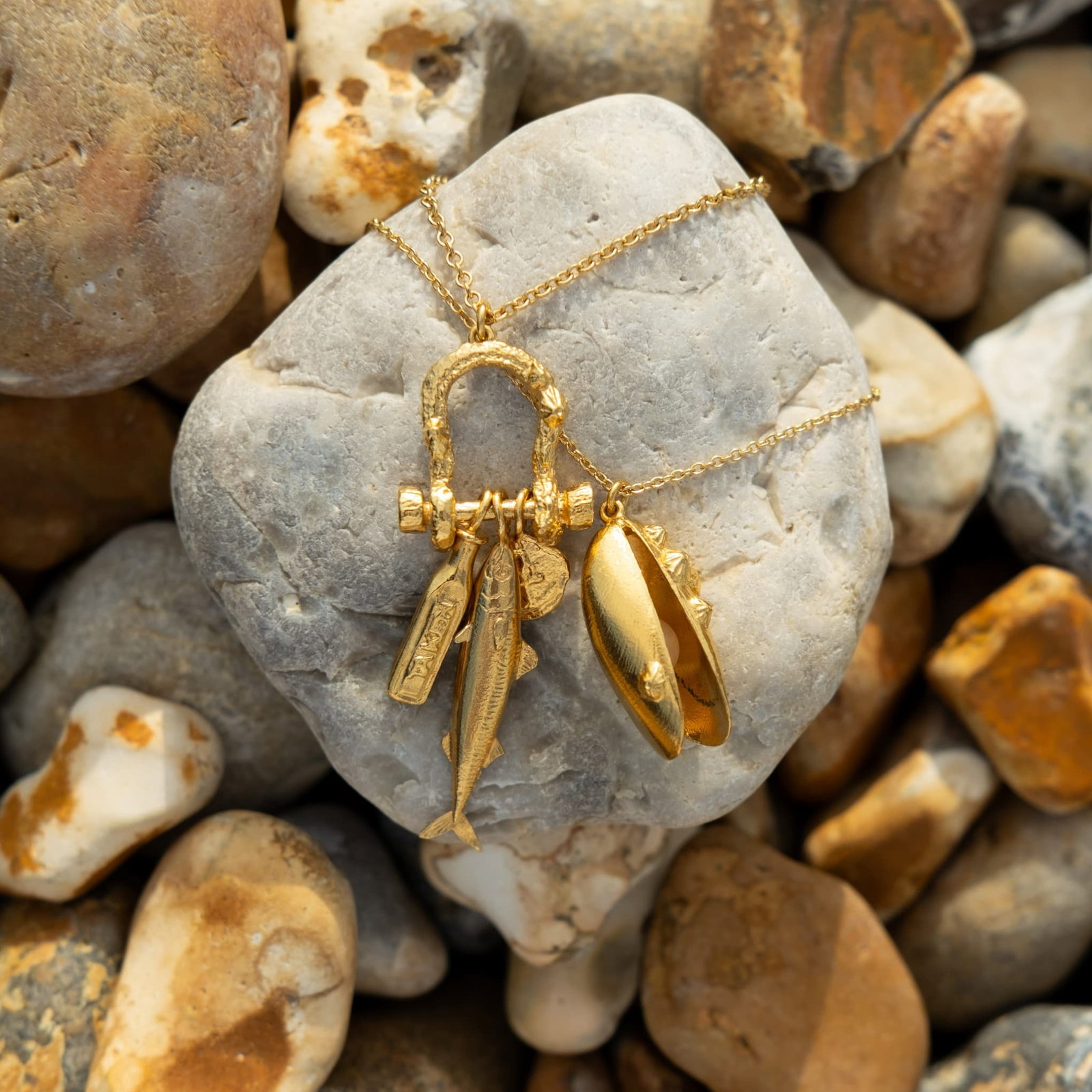 Engraved Shell Necklace in sterling silver displayed on a smooth pebble, with a natural background of rounded beach stones in warm neutral tones.