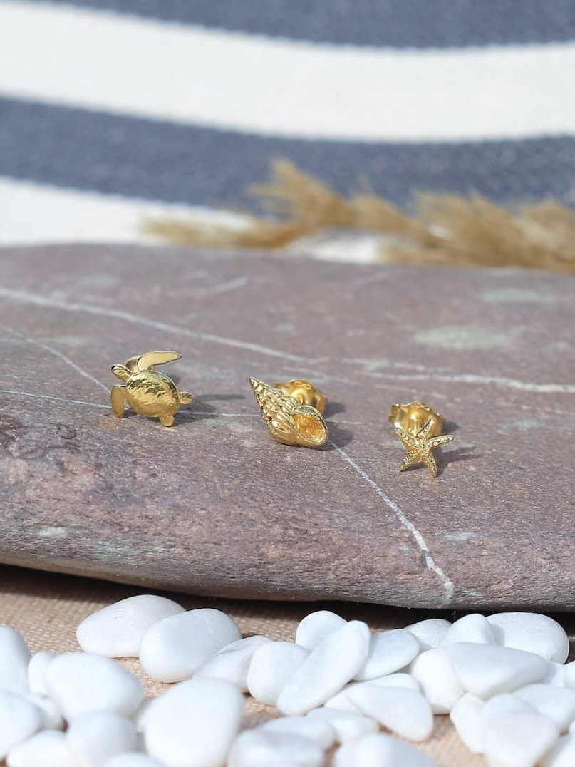 Teeny Tiny Collection gold stud earrings featuring miniature sea-inspired designs including a turtle, shell and starfish, displayed on a smooth beach stone with white pebbles in a coastal setting.