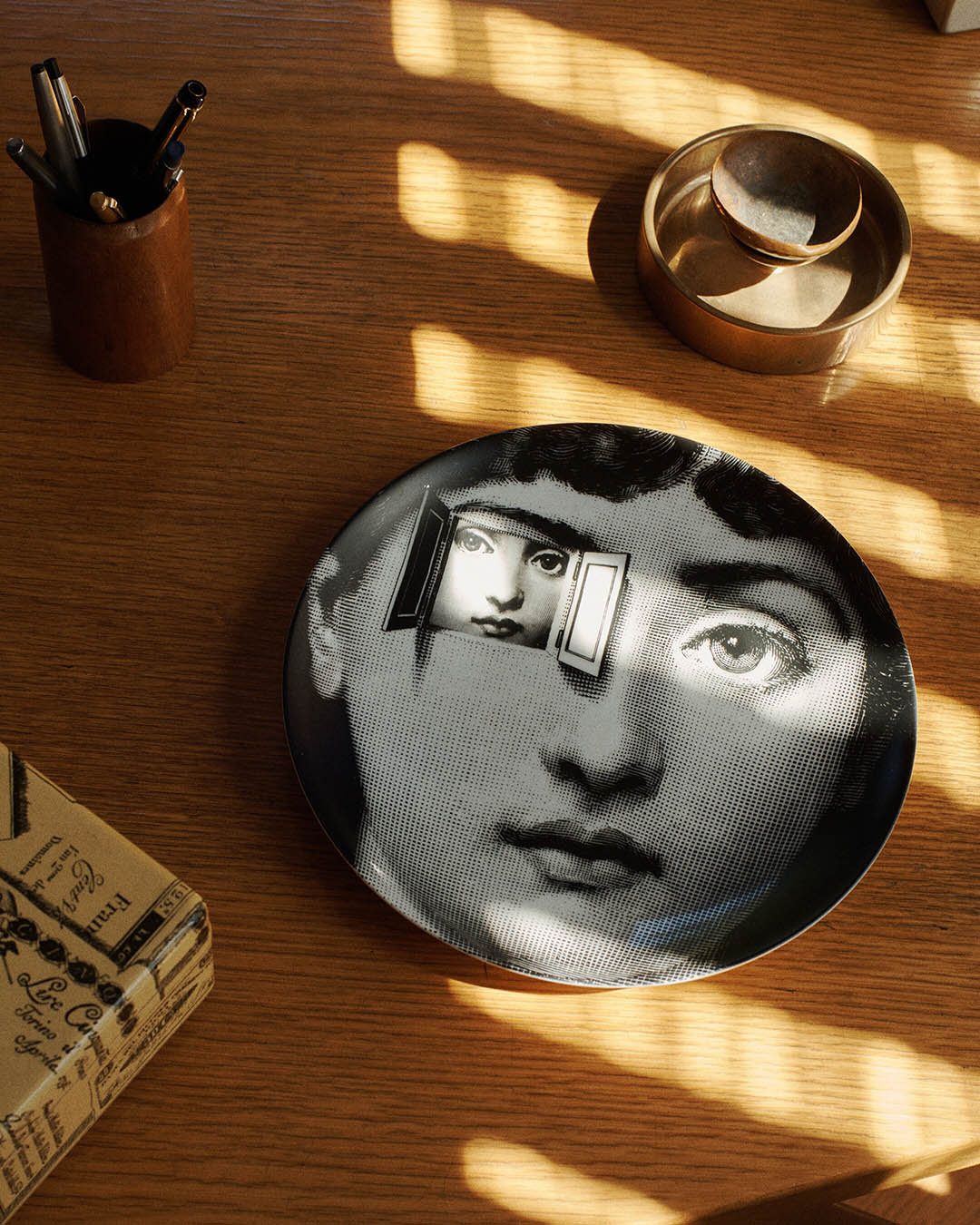 Sunlit wooden desk with decorative black-and-white porcelain plate showing a stylised female face, brass dishes and pens