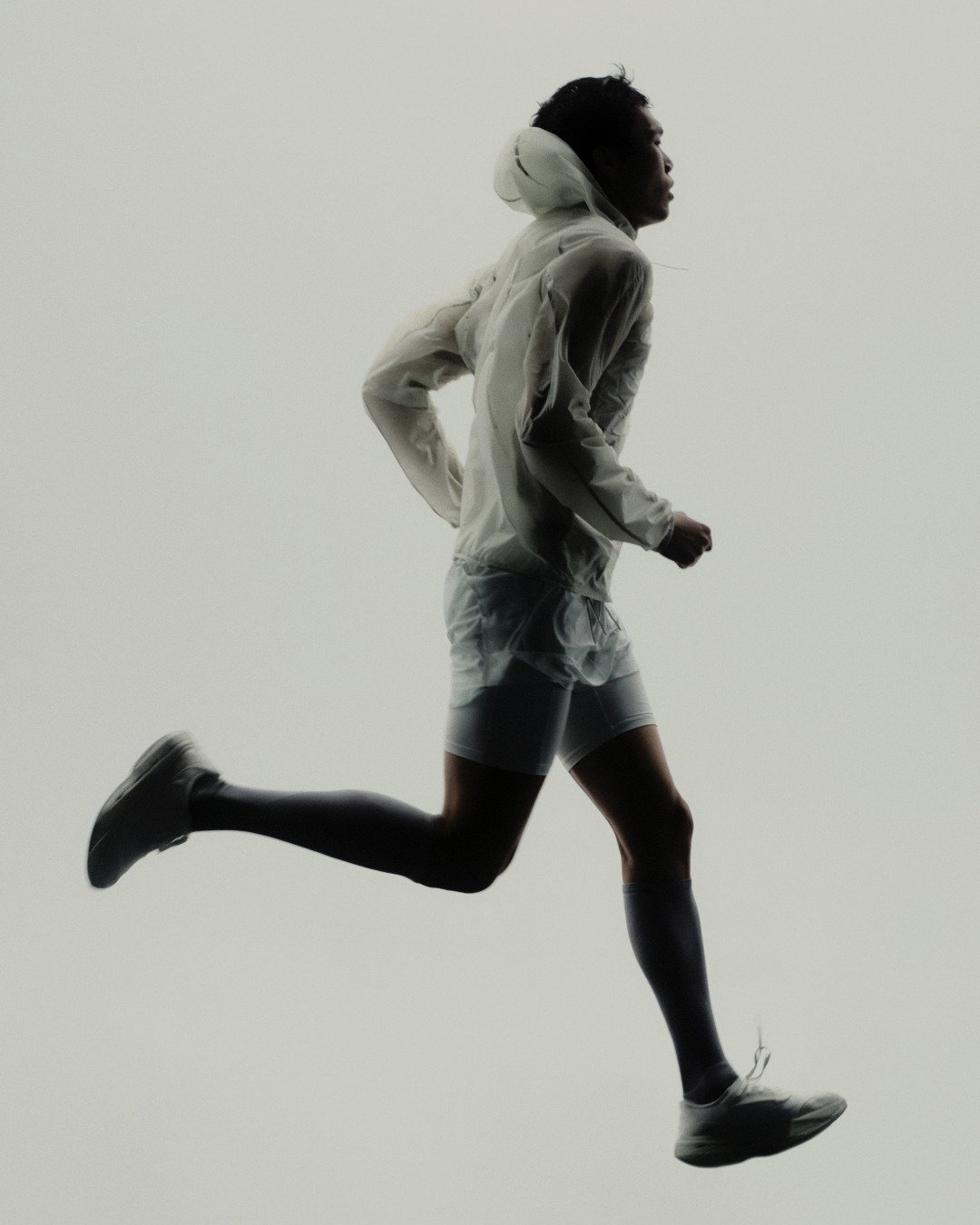Runner captured mid-stride wearing a translucent rain jacket, shorts and trainers against a pale minimalist background