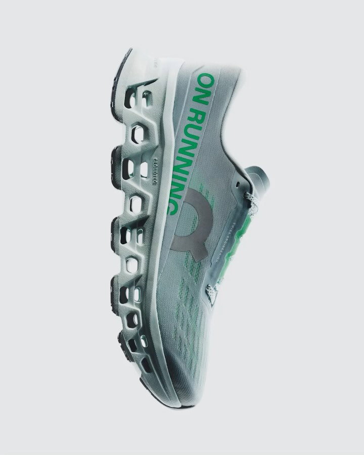 Side view of grey On Running athletic shoe with green "ON RUNNING" text and distinctive hollow cushioned sole