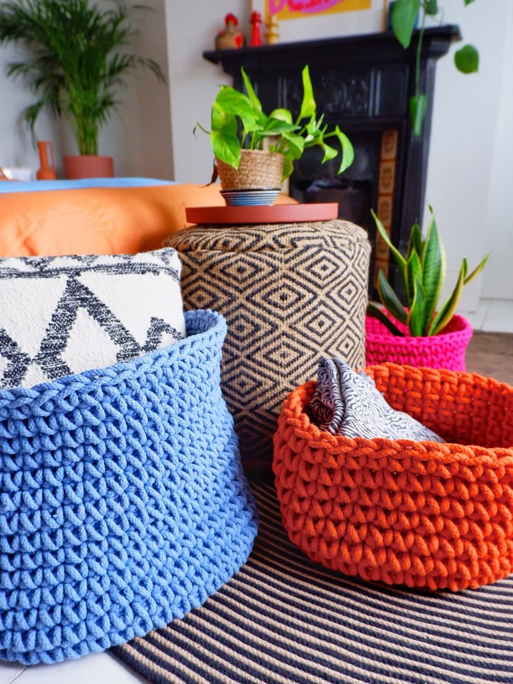 Brightly colored Homescapes woven baskets in blue, orange, and pink styled with a geometric pouffe and indoor plants.