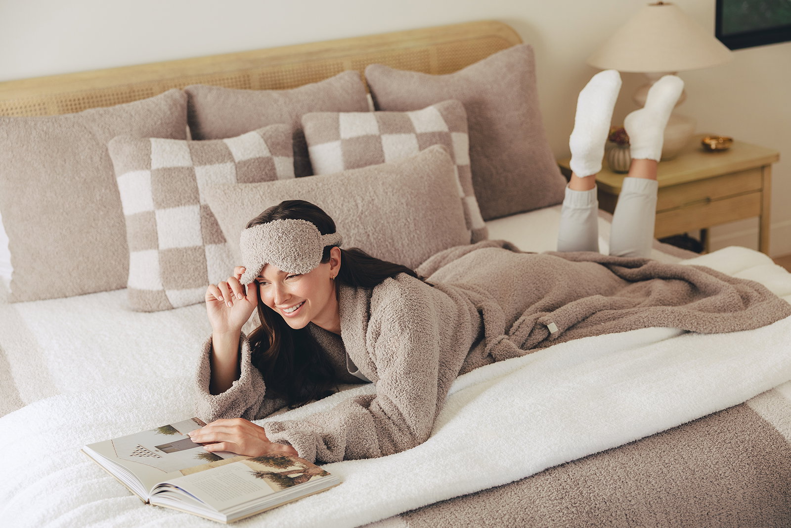 Woman relaxing on a bed in a plush robe with a matching eye mask, surrounded by soft pillows and a cozy throw blanket while reading a book.