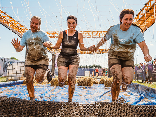 three women smiling and holding hands make their way through a muddy outdoor obstacle course.