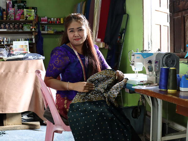 a woman is sat at a sewing table. She has a tape measure around her neck and is holding a patterned piece of fabric. 