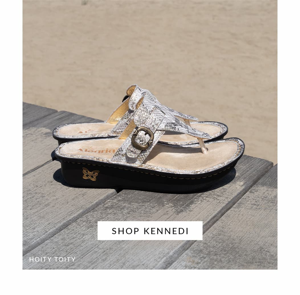 Alegria Kennedi thong sandals in Hoity Toity print featuring a snakeskin-inspired leather upper, adjustable buckle, and signature Classic Footbed, displayed on a wooden boardwalk by the sandy beach.