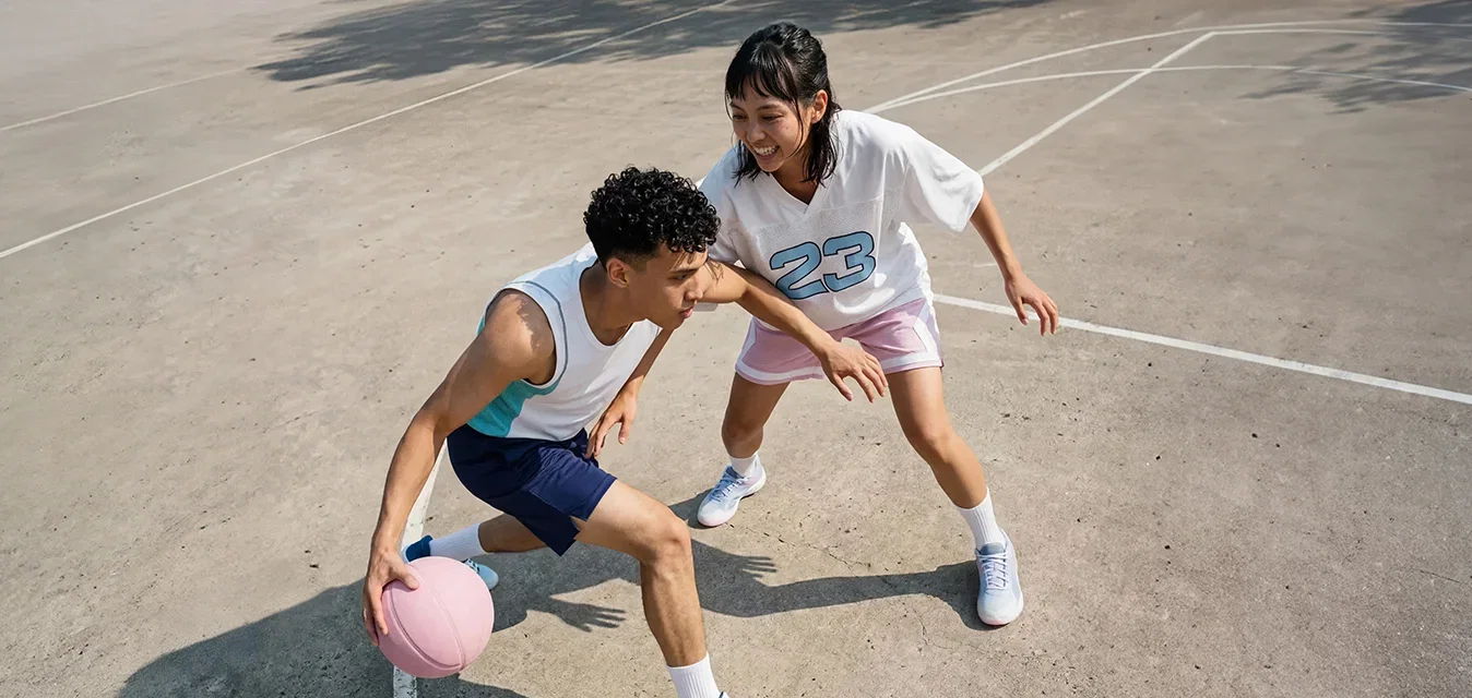 Un homme fait des dribbles avec un ballon de basket rose tandis qu'une femme en maillot numéro 23 le défend sur un terrain extérieur.