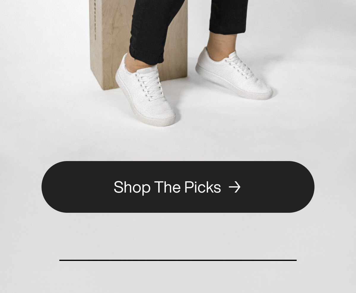 "White sneakers on a person sitting, with a "Shop The Picks" button below. Simple, stylish footwear display."