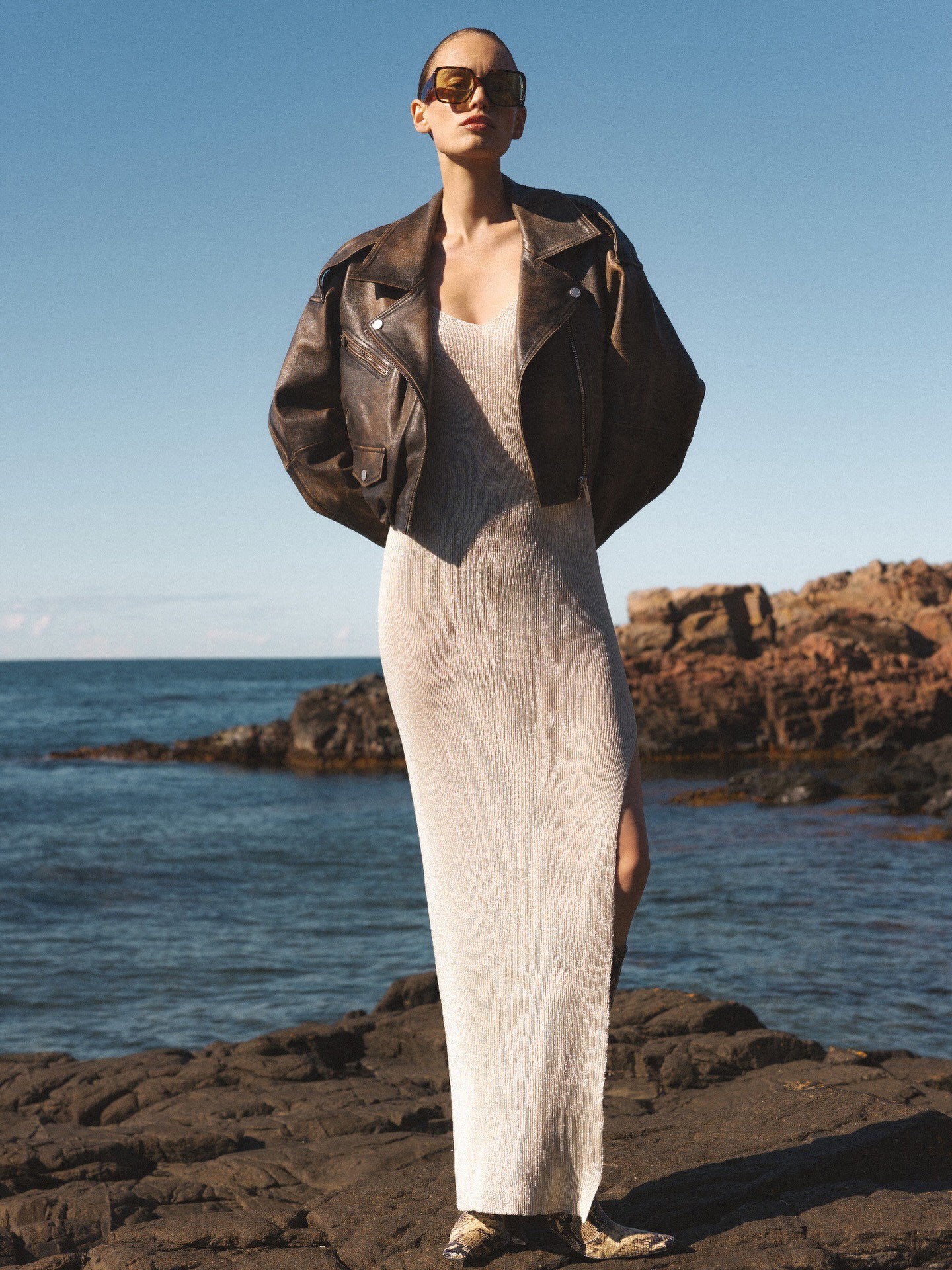 Model wearing a cream knit maxi dress, oversized brown leather jacket and sunglasses standing on a rocky shoreline by the sea