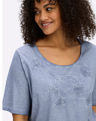 Model in blauem Shirt in Oil-washed-Optik mit Blütenstickerei vorn.
