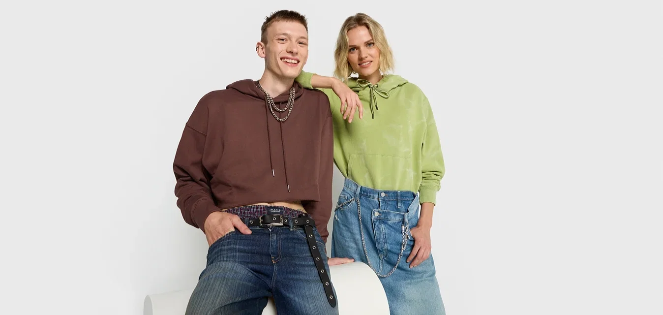 Dos modelos posan juntos, uno con una sudadera marrón y el otro con una camiseta verde, ambos vistiendo denim. Las características incluyen un corte holgado y texturas variadas.