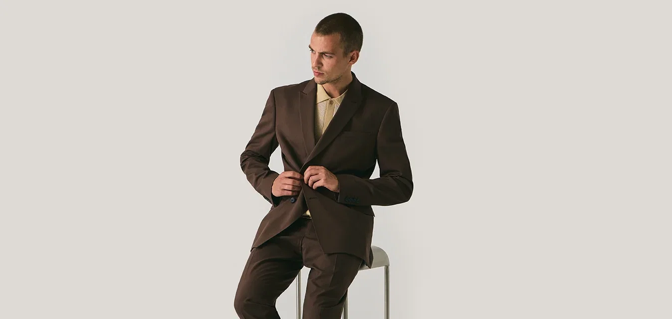 Traje marrón con chaqueta ajustada y piernas rectas, combinado con una camisa de tono claro y zapatos marrones, sentado en un taburete de metal.