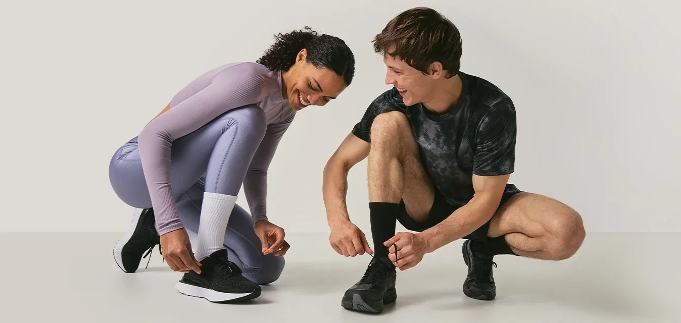 Dos personas con ropa deportiva se agachan para atarse las zapatillas de correr sobre un fondo liso de color claro.