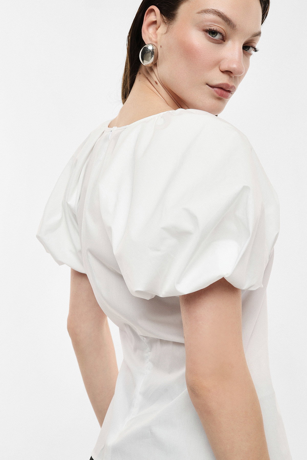 Woman wearing white puff-sleeve blouse and large silver disc earring, looking over her shoulder against a plain background