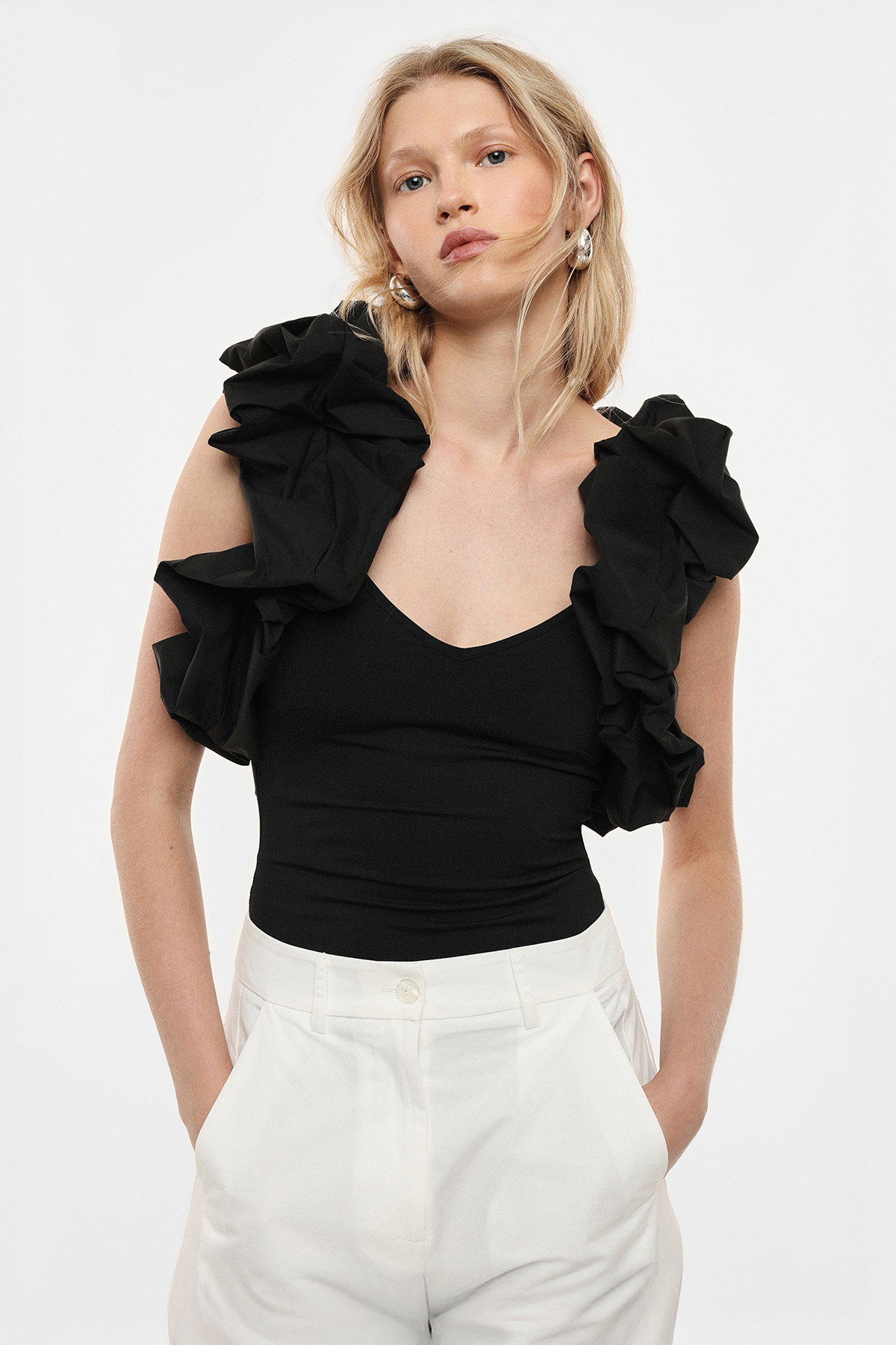 Woman wearing black ruffle-shoulder top and white trousers, hands in pockets, fashion portrait on plain background