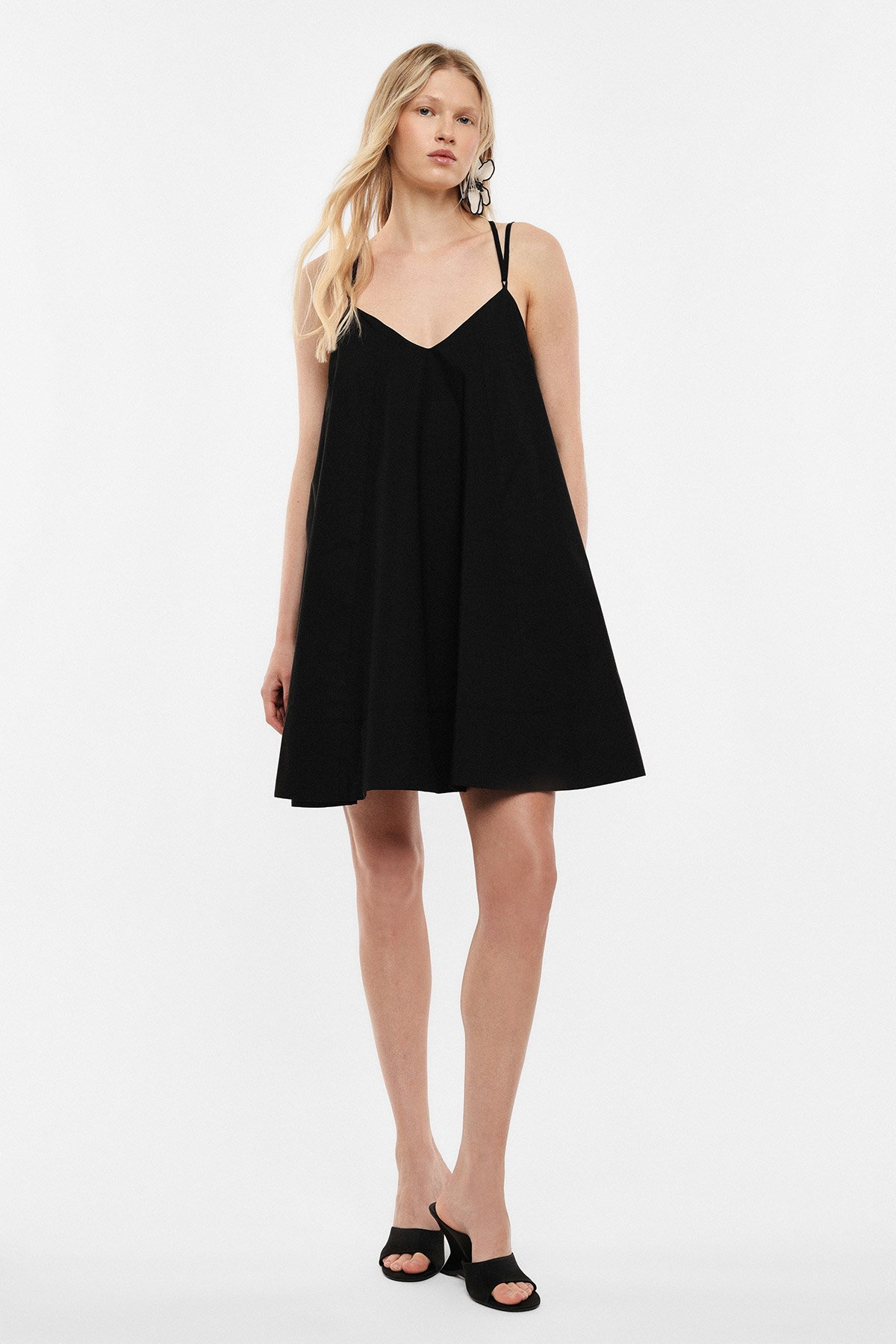 Woman wearing black spaghetti‑strap A-line mini dress with black heeled mules and statement earrings on white background