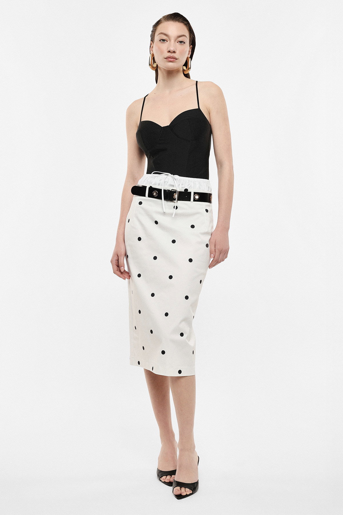 Female model wearing black spaghetti‑strap bodysuit with white high‑waist polka‑dot pencil skirt, black belted waist and black open‑toe heels