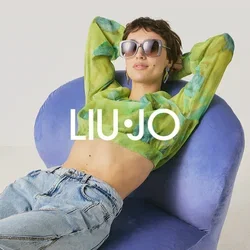 Persona con occhiali da sole, top corto verde a motivo e jeans che si rilassa su una sedia moderna viola contro uno sfondo chiaro e uniforme.