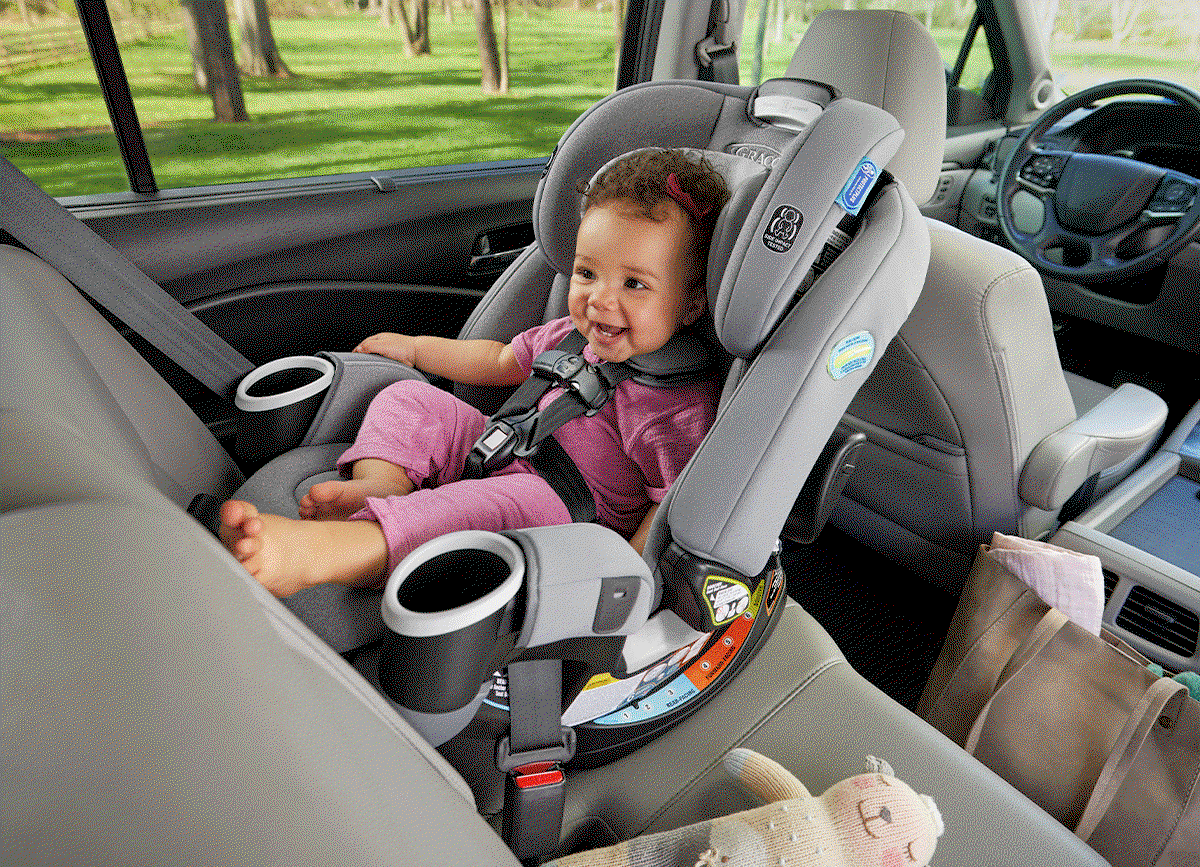 Smiling baby sitting in a gray car seat in the back seat of a vehicle, surrounded by bags and toys.