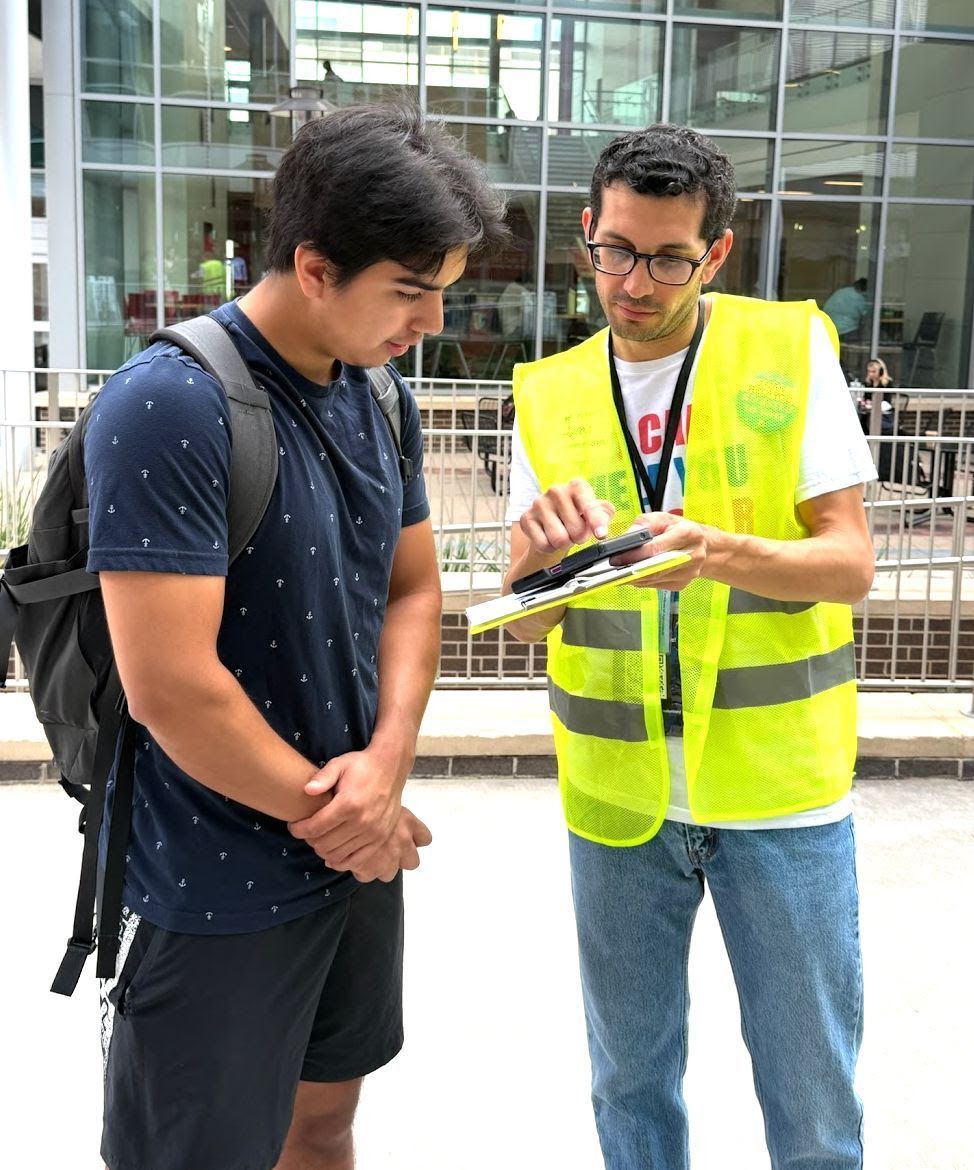 Image of a volunteer wearing a high-visibility vest (right) helping someone check their voter registration (left), showing them a clipboard and something on his phone