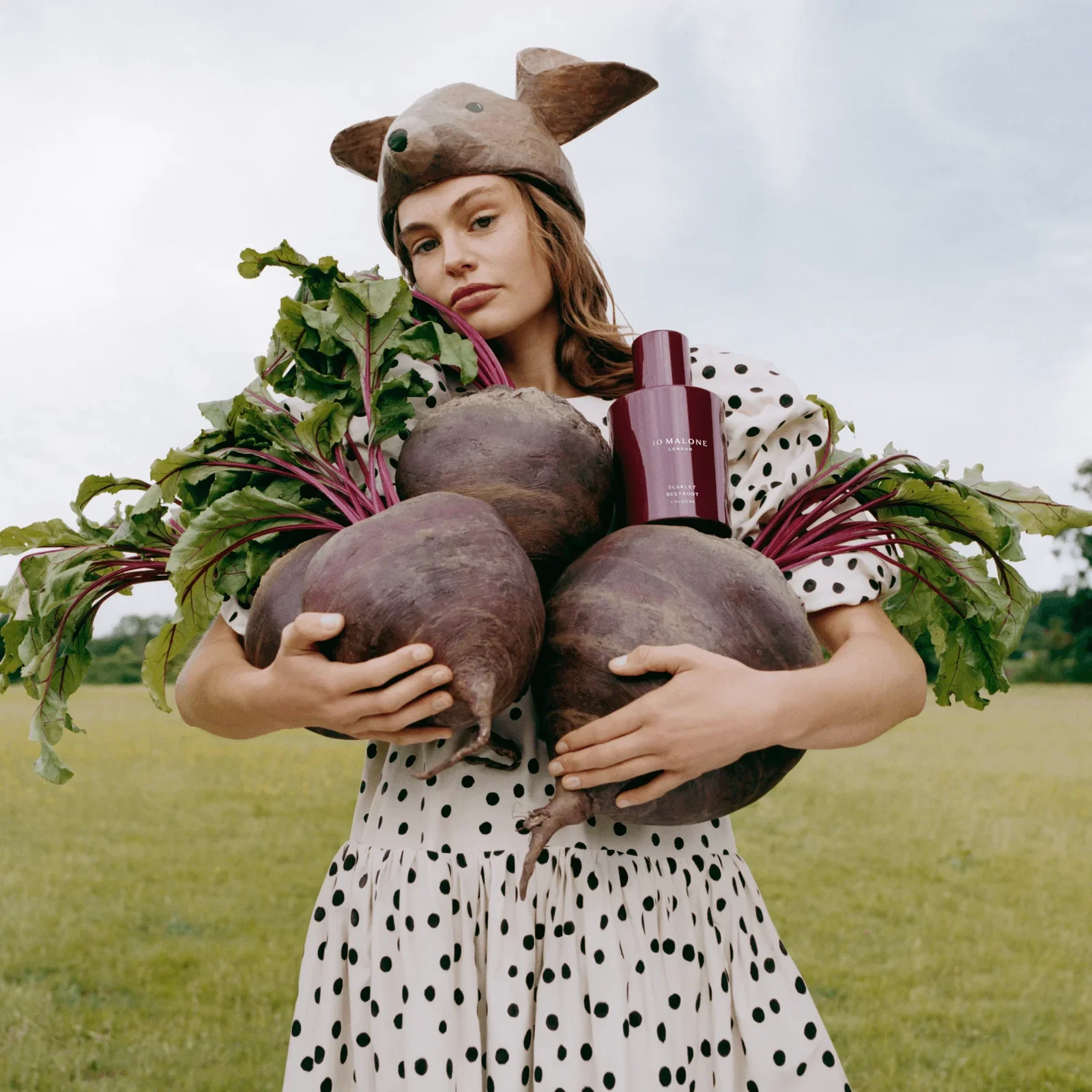 Woman holding four giant beetroots and a bottle of Jo Malone Scarlet Beetroot Cologne, wearing an animal hat and polka-dot clothing in the countryside.