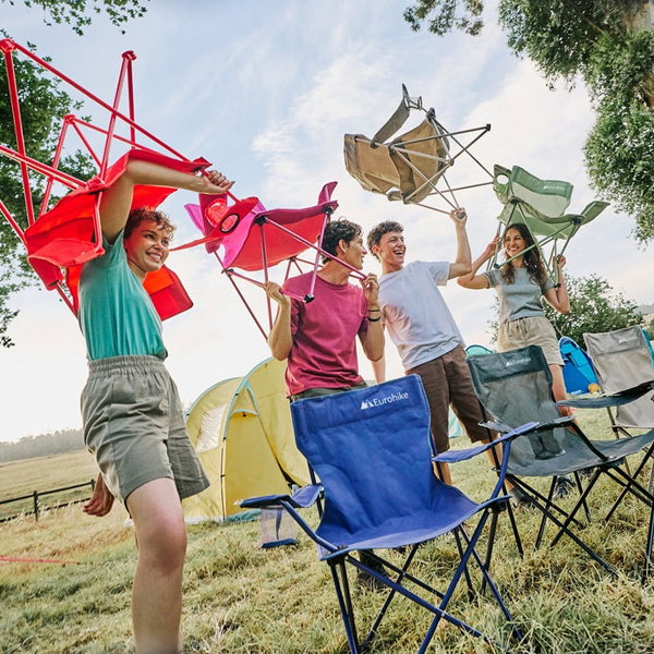 A person sets up a reclining camping chair in front of a large tent on a grassy campsite. Additional folding chairs and camping gear are placed nearby, with trees and hills visible in the background.