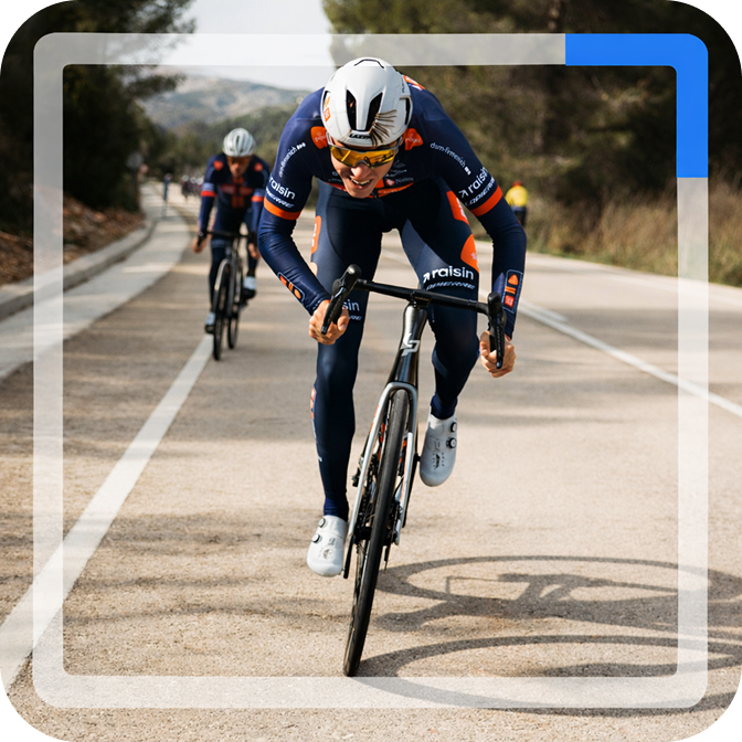 Un ciclista profesional con equipación azul marino y naranja avanza concentrado en una posición aerodinámica por una carretera, seguido de cerca por otro compañero de equipo.