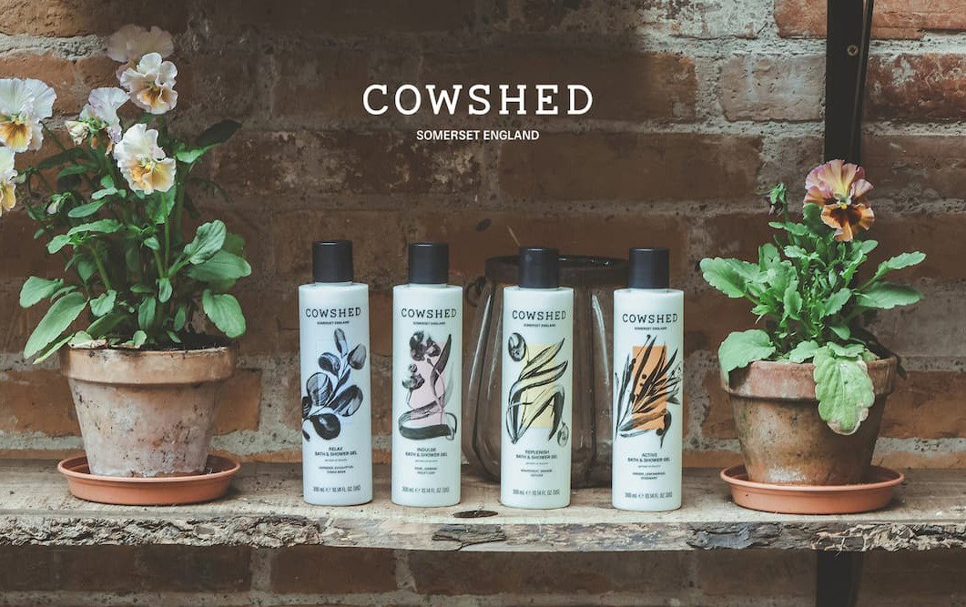 Four bottles of Cowshed bath & shower gel on a shelf with potted flowers either side