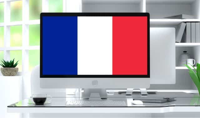 A computer monitor on a modern desk displays the French flag. There are office supplies, a plant, and a cup of coffee on the desk.