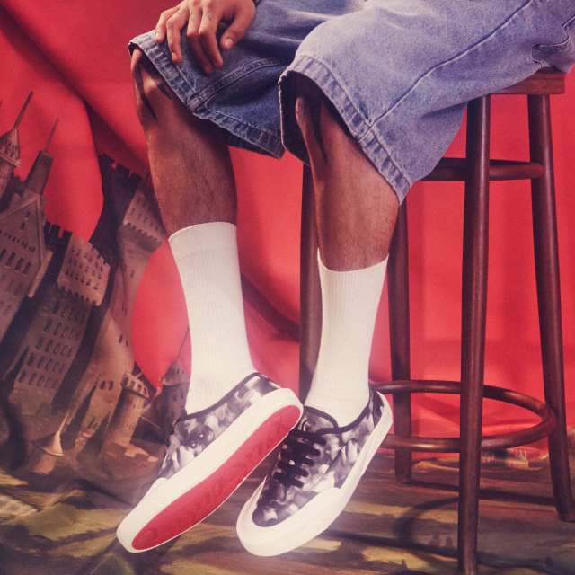 Person sitting on a stool, wearing denim shorts, white socks, and patterned sneakers with red soles, against a red background with illustrations.