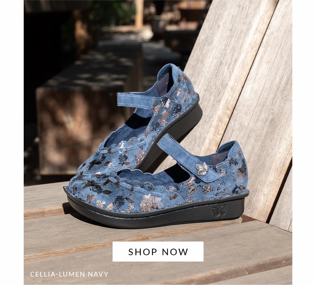 A side profile image of Cellia Lumen Navy mary jane shoe with adjustable hook-and-loop strap. Shown resting against a wooden deck.