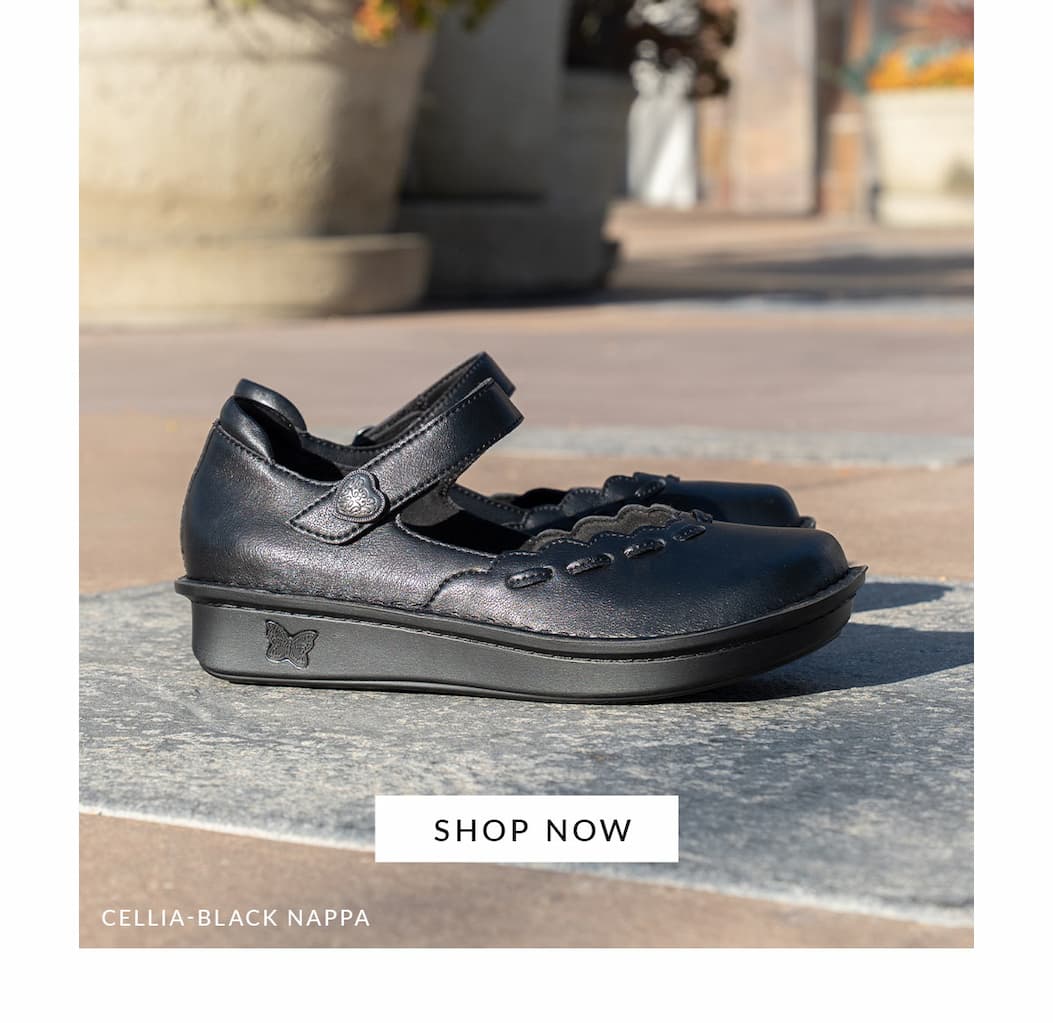 Cellia Black Nappa mary jane with hook-and-look adjustable strap with decorative scalloped edging. Shown placed in a courtyard on top of stamped concrete.