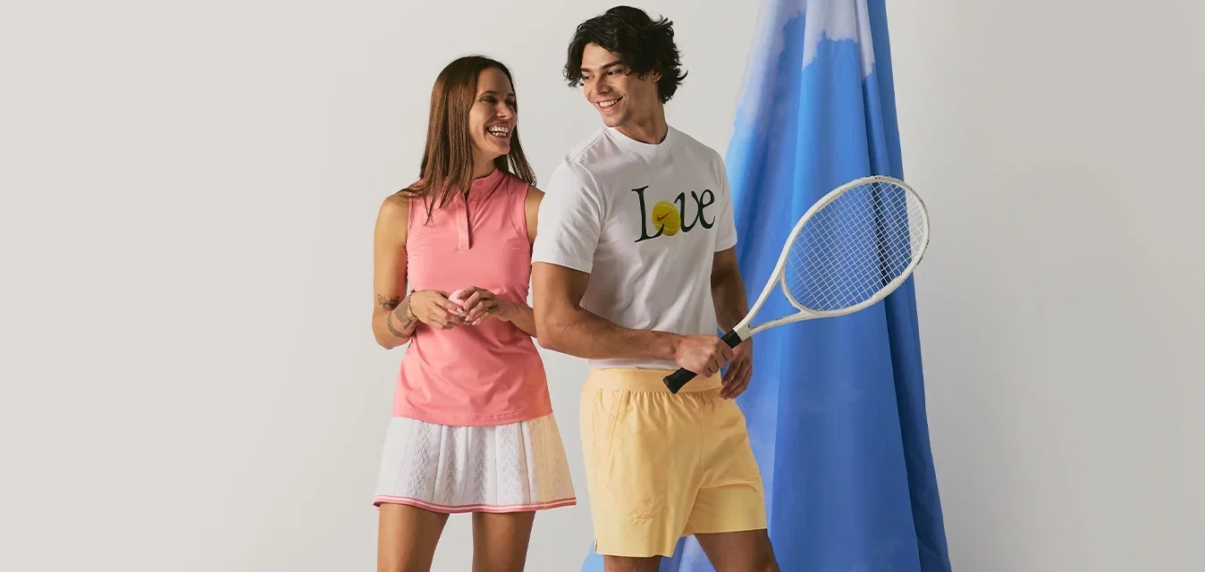 Un hombre y una mujer sonrientes con ropa de tenis están junto a un fondo de tela azul, con el hombre sosteniendo una raqueta de tenis.