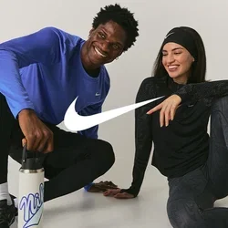 Dos personas con ropa deportiva sentadas en el suelo, sonriendo; una sostiene una botella de agua, ambas parecen relajadas y amistosas.
