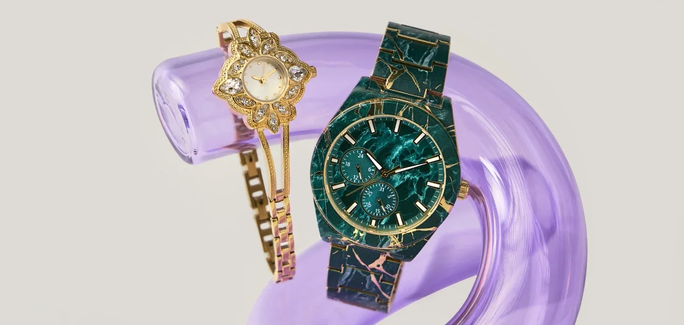 Reloj de pulsera con flores doradas y reloj de muñeca con patrón de mármol verde oscuro descansando sobre un soporte curvado translúcido púrpura contra un fondo claro.