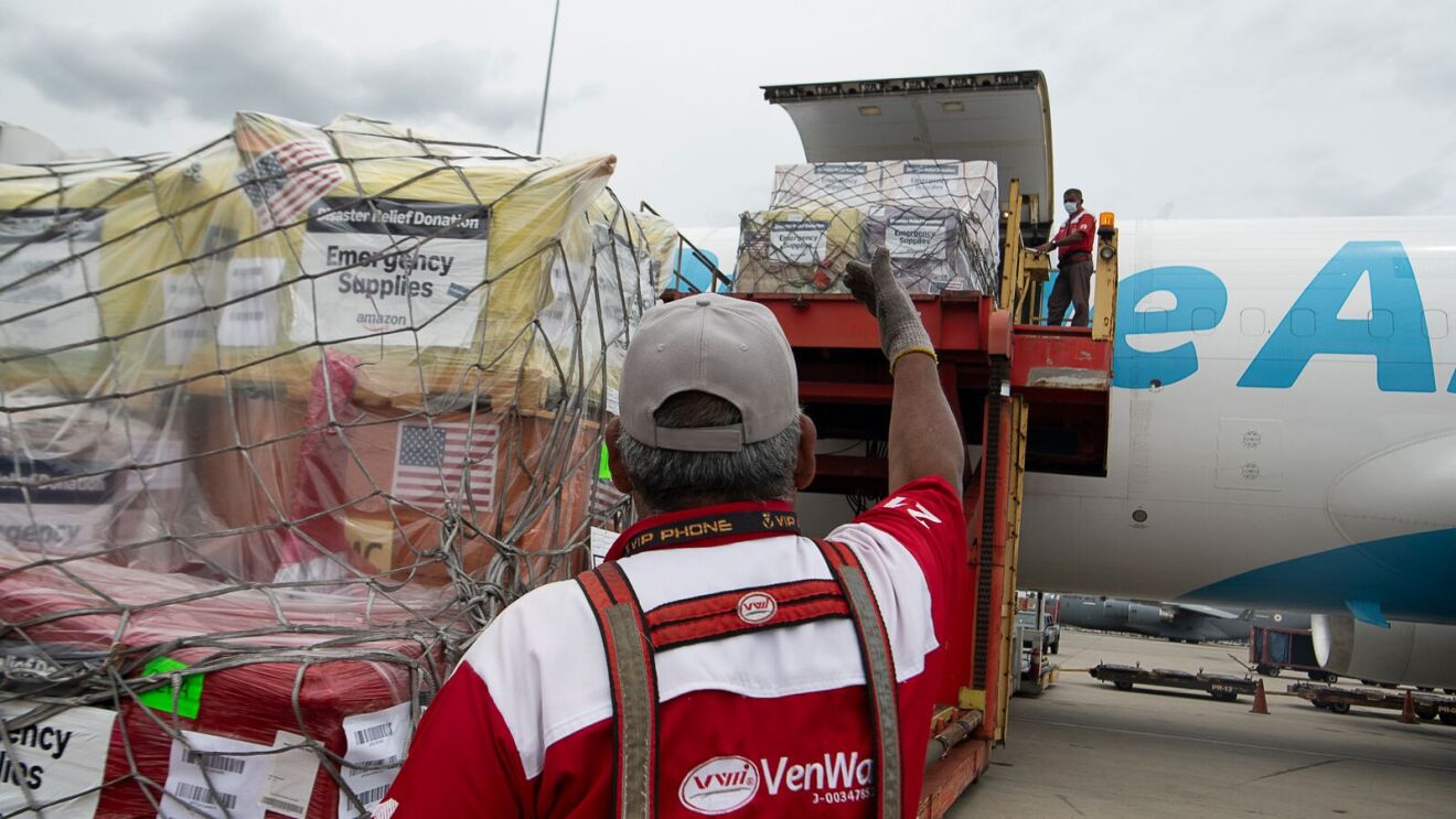 Emergency relief supplies from Amazon, including packages with US flags, are being loaded by workers onto a large airplane.