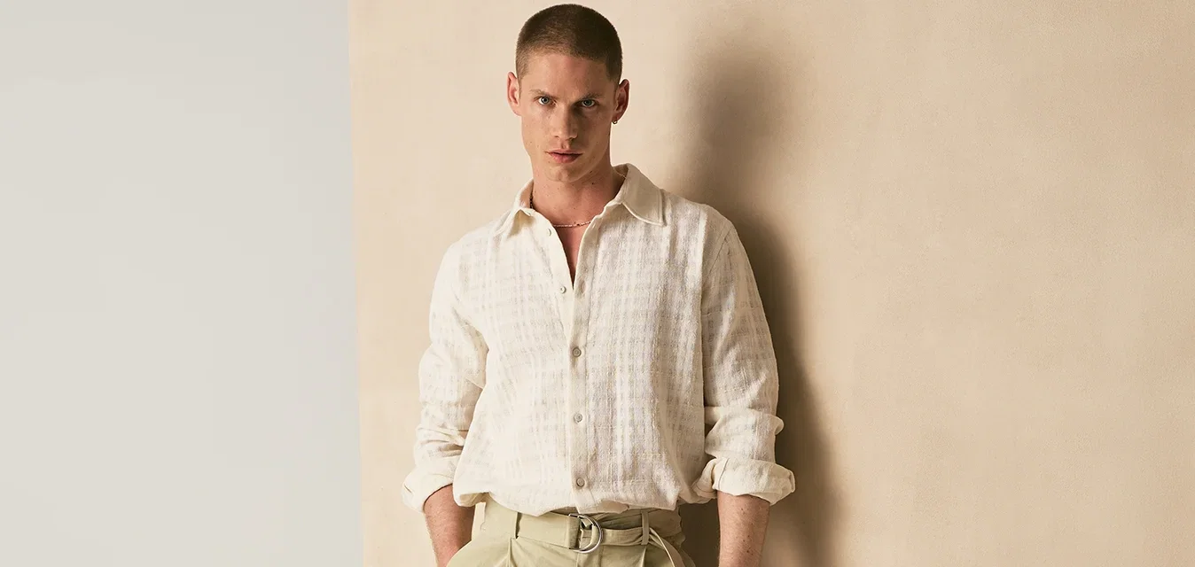 Man in beige wijde broek en wit gestructureerd hemd staat met handen in de zakken tegen een tweekleurige beige en gebroken witte muur.