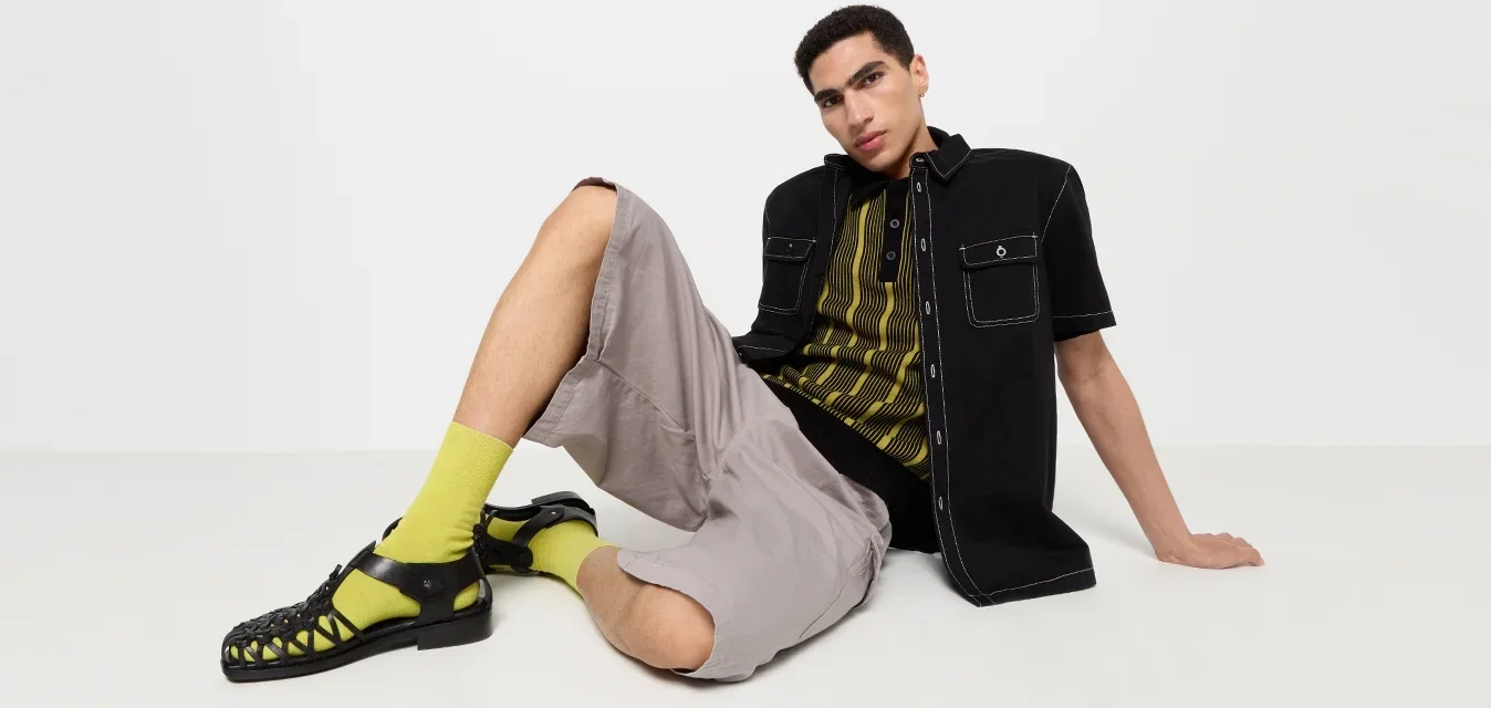 Zwarte korte mouwen shirt met zakken, gele gestreepte t-shirt eronder, grijze shorts, felgroene sokken en zwarte sandalen met riemen.