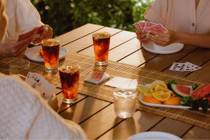 Friends playing cards around an outdoor table with iced drinks and fresh fruit