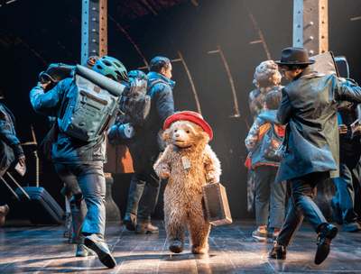 A small bear in a red hat and suitcase stands amidst a bustling crowd on stage.