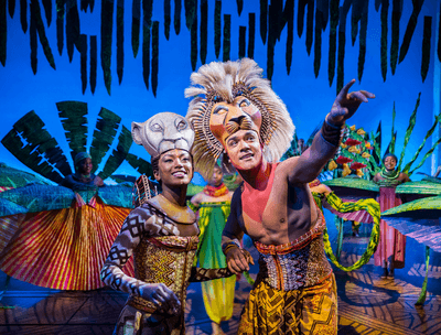 A vibrant stage performance featuring two characters in elaborate costumes, surrounded by dancers.