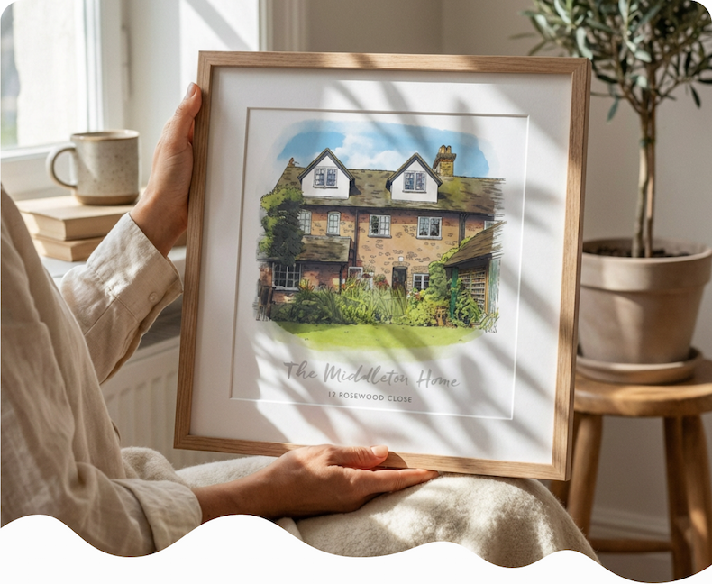 A framed watercolour illustration of a stone cottage personalised with the family name, held up by someone in a linen shirt beside a window with a plant in the background