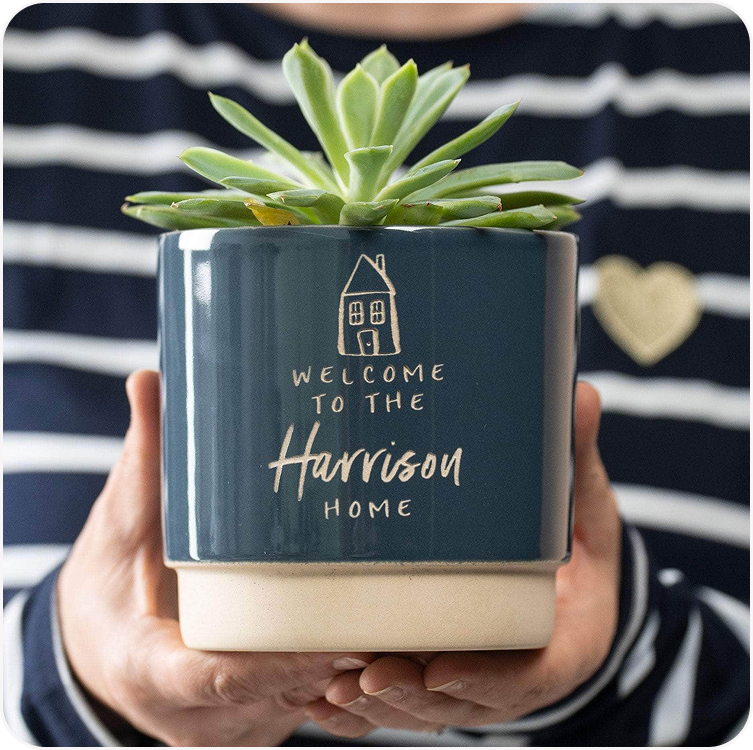 A dark ceramic plant pot engraved with "Welcome to the Harrison Home" held against a striped top, planted with a succulent