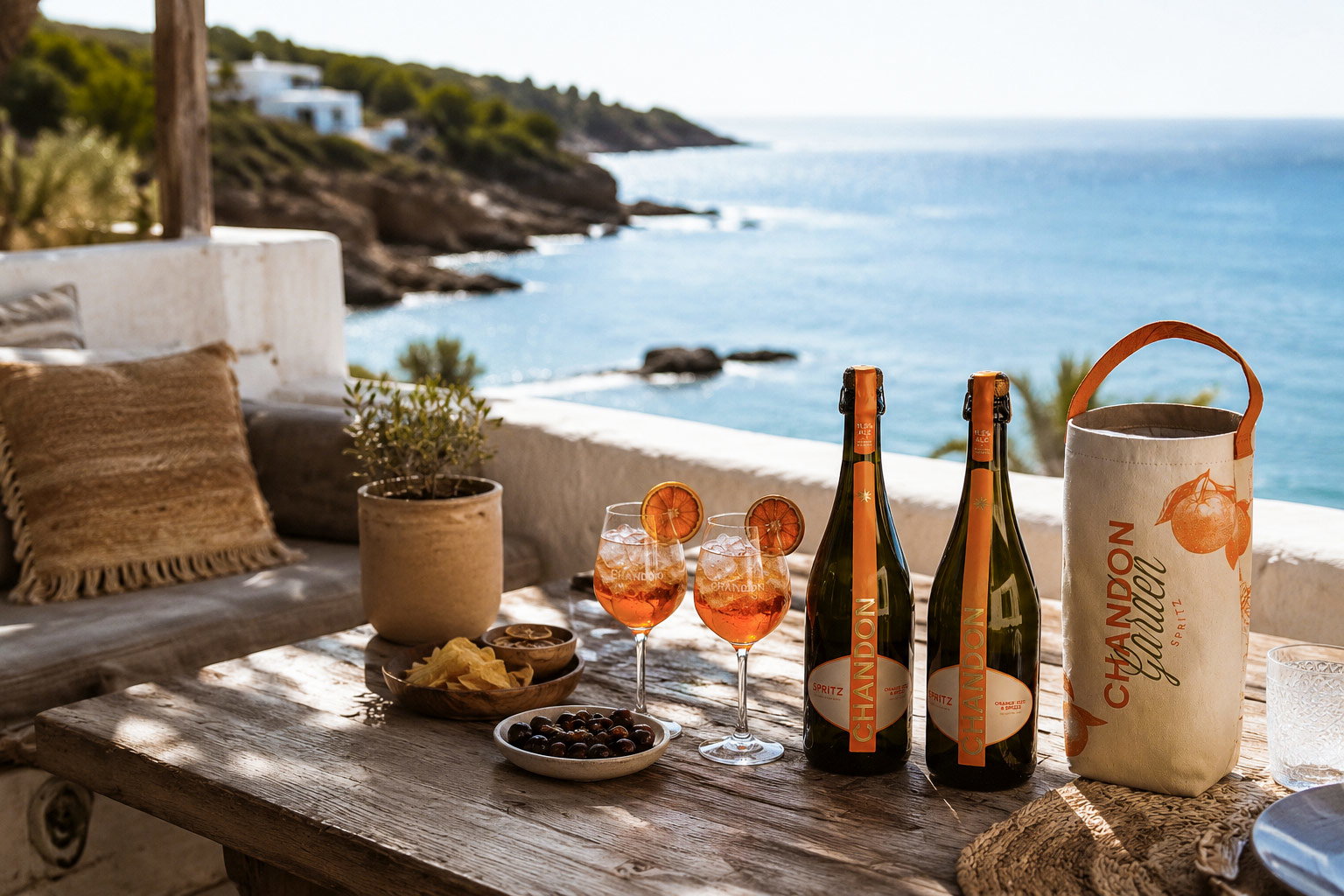 Seaside terrace table with two chilled Aperol spritzes, two Chandon bottles, snacks and sunlit Mediterranean bay view